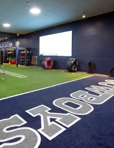 Athletic training facility featuring high-quality turf surface with "COWBOYS" branding, agility training area, weights, and fitness equipment, emphasizing performance and safety for athletes.