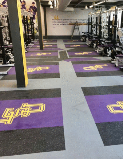 Weight room featuring high-performance rubber flooring with custom purple and yellow logos, showcasing team identity, designed for safety and sound absorption, surrounded by weightlifting equipment.