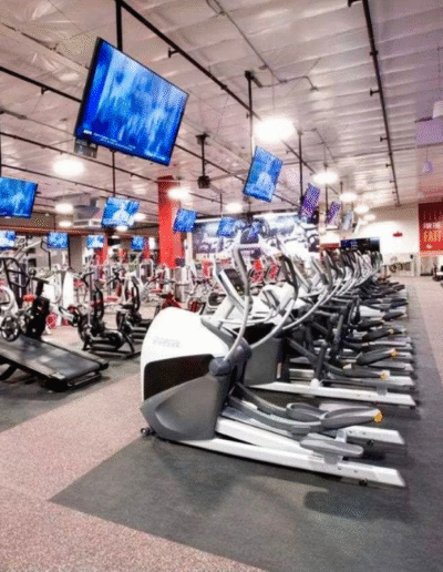 Gym interior featuring multiple elliptical machines on rubber flooring, with large screens displaying workout content, emphasizing a high-performance fitness environment.