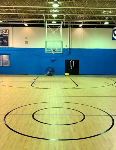 Basketball court featuring polished hardwood flooring, black court markings, and a blue wall backdrop, demonstrating refinished sports flooring by Sport Pro Surfacing.