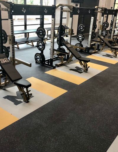 Weight room with custom rubber flooring featuring team logos, benches, and weightlifting equipment, designed for safety and performance.