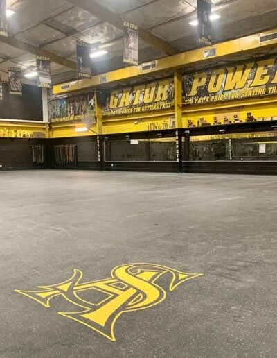 Flooring in sports facility featuring a prominent yellow and black logo with "S" and "H" design, surrounded by motivational banners and trophies, showcasing custom branding by Sport Pro Surfacing.