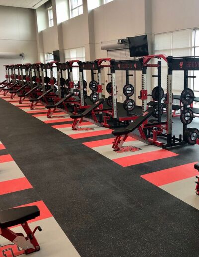 Weight room featuring custom-designed rubber flooring with embedded logos, multiple weightlifting stations, and a cohesive red and gray color scheme, emphasizing safety and team identity for athletic training.