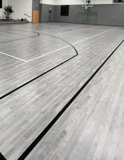 Newly refinished basketball court showcasing smooth gray hardwood flooring with black court lines and markings, emphasizing high-quality sports flooring by Sport Pro Surfacing.
