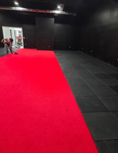 Athletic facility interior showing red performance turf installation alongside black rubber flooring, with a worker preparing the surface for training activities.