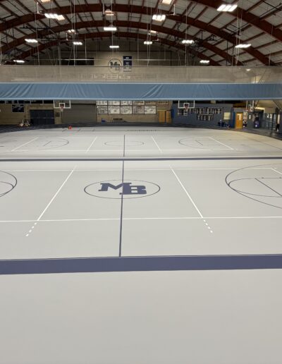 Basketball court featuring a gray surface with blue lines and an "MB" logo at the center, showcasing refinished flooring for optimal playability and aesthetics.