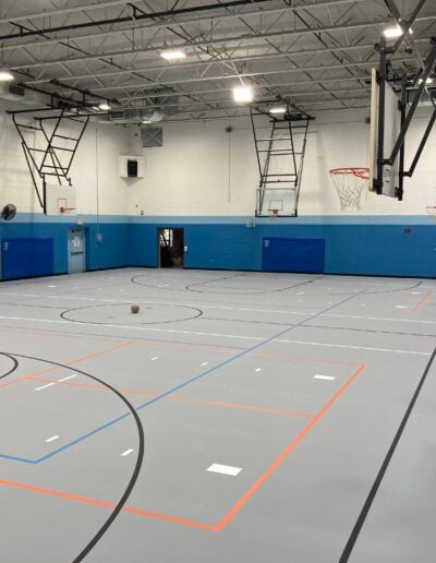 Basketball court with gray flooring, blue walls, and basketball hoops, showcasing refinished sports flooring by Sport Pro Surfacing.