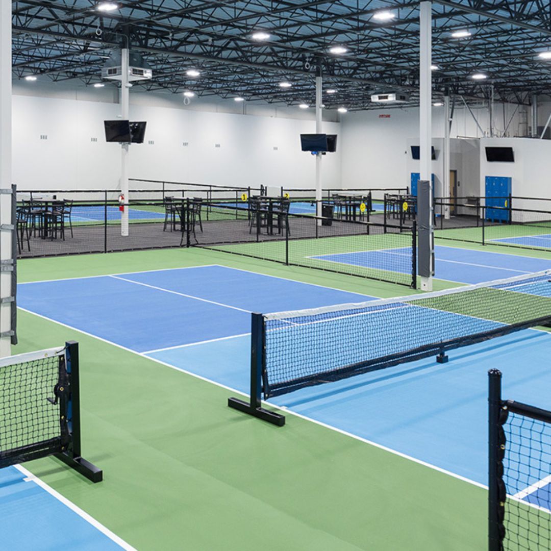 Indoor pickleball courts featuring blue and green surfaces, nets, and seating areas, showcasing a modern athletic facility designed for sports activities.