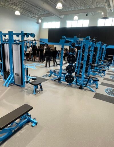 Gym interior featuring blue and black weight training equipment, rubber flooring, and a group of people gathered, highlighting a functional fitness area designed for performance and safety.