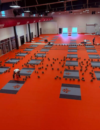 Rubber flooring installation in a weight room, featuring custom logo designs and vibrant orange surfaces, with workers arranging flooring tiles for enhanced safety and team identity.