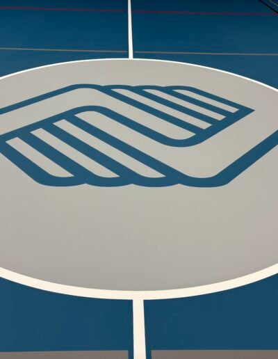 Logo of a sports facility featuring a stylized hand design, prominently displayed on a blue and gray athletic floor surface, representing custom branding solutions by Sport Pro Surfacing.